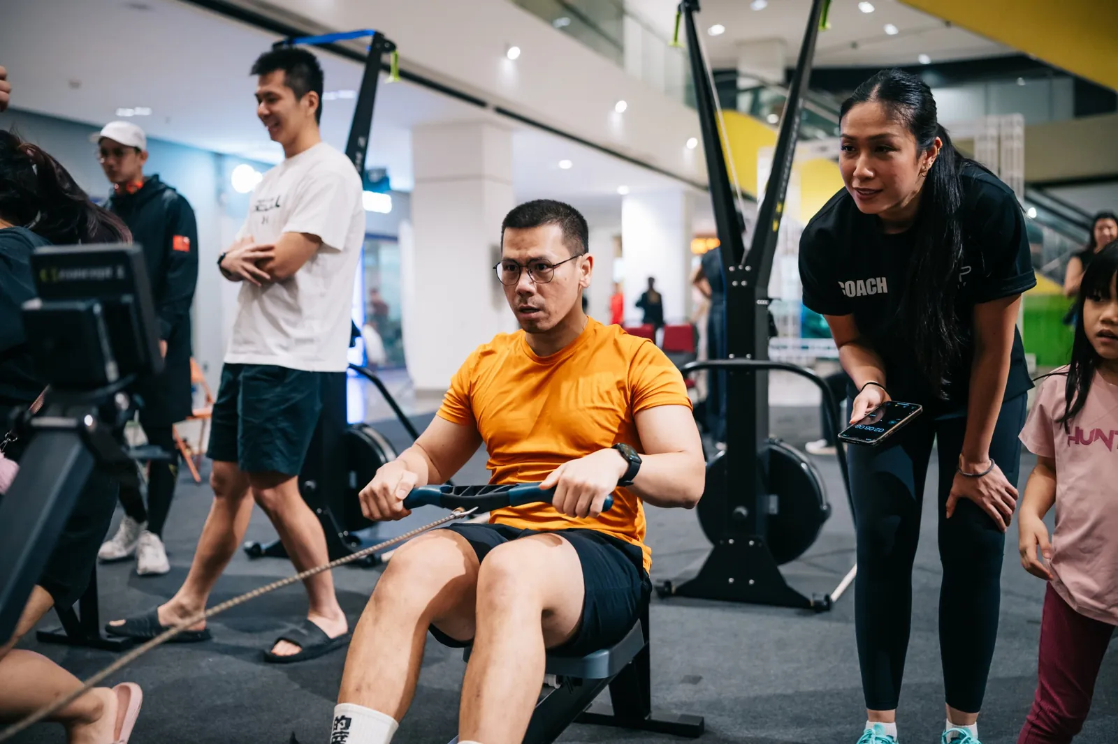 A rower in orange, a coach with a stopwatch, kids watching